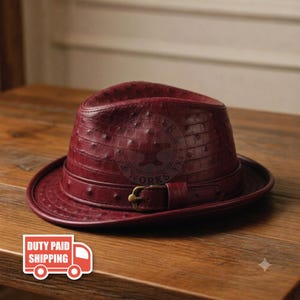 May include: A deep burgundy fedora hat featuring a textured ostrich leather pattern. The hat includes a matching leather band around the crown, secured with a small, metallic gold-toned buckle. The surface has a distinct quill-dot texture.