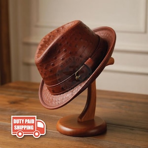 May include: A brown fedora hat with a textured ostrich-skin pattern, featuring a matching leather band around the crown and a small metallic buckle detail on the side. The hat is displayed on a curved wooden stand, showcasing its structured brim.
