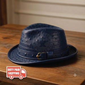 May include: A deep blue fedora hat with a textured ostrich-like pattern, featuring a matching blue hat band with a brass-toned metal buckle. A circular logo with the text "LEATHER WORKS EST" is embossed on the front of the crown.
