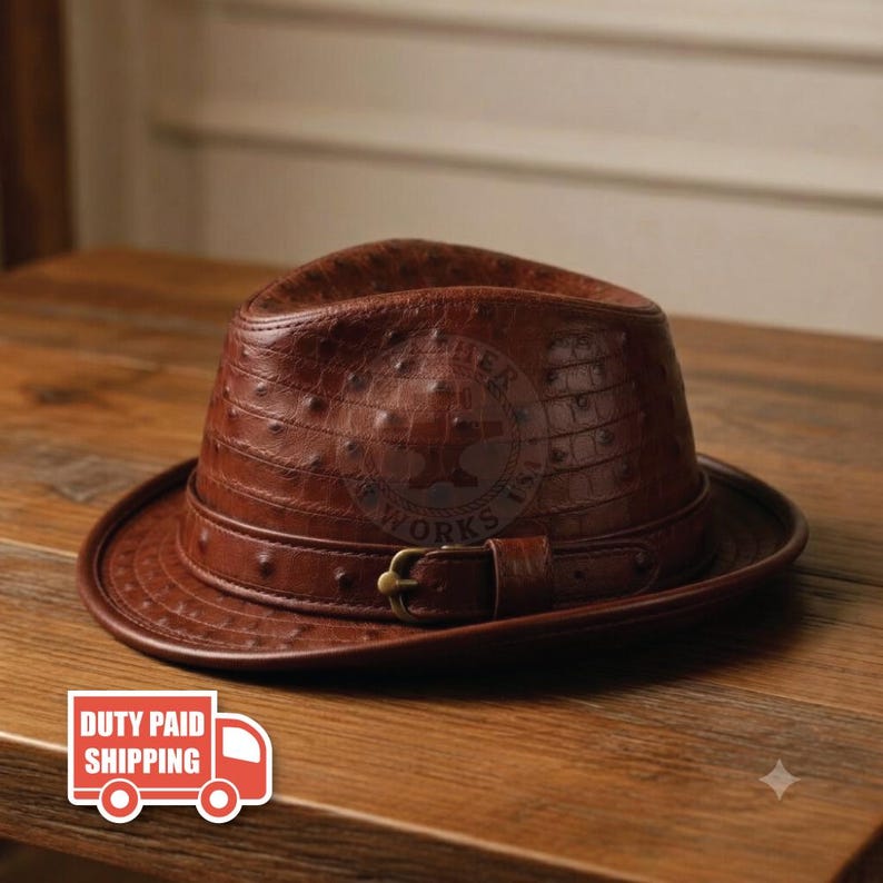 May include: A brown fedora hat featuring a textured ostrich-skin pattern, a matching leather band, and a brass-toned metal buckle. The hat has a structured crown and a curved brim, showcasing a detailed embossed logo on the front surface.