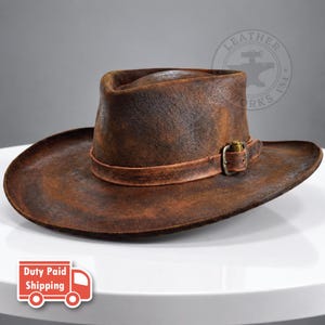 Puede incluir: Un sombrero de vaquero de cuero marrón con un acabado texturizado y desgastado. El sombrero presenta una banda de cuero a juego alrededor de la copa, asegurada por una hebilla metálica con acabado en tono latón, creando un aspecto robusto.