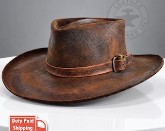 May include: A distressed brown leather cowboy hat with a textured, weathered finish. The hat features a matching leather band around the crown, secured by a metallic buckle with a brass-toned finish, creating a rugged and worn appearance.