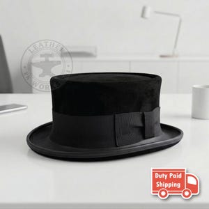 Puede incluir: Un sombrero de copa negro con una corona plana y un ala ancha. El sombrero está adornado con una cinta de grosgrain negra que envuelve la base de la corona, con un pequeño detalle de lazo en el lateral. La textura parece lisa y mate.