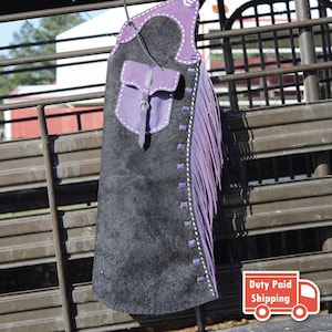 May include: A black leather chaps piece featuring purple leather trim, a matching purple pocket with a buckle closure, and long purple fringe along the side. The garment is accented with white stitching and square cutouts along the edge.