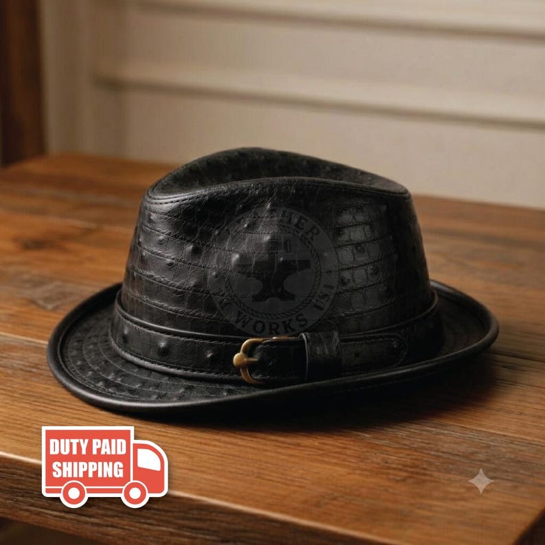 May include: A black fedora hat with a textured ostrich-like pattern, featuring a matching black hat band with a metallic buckle. The front of the hat displays a circular logo with an airplane silhouette and the text "ARCHER WORKS USA".