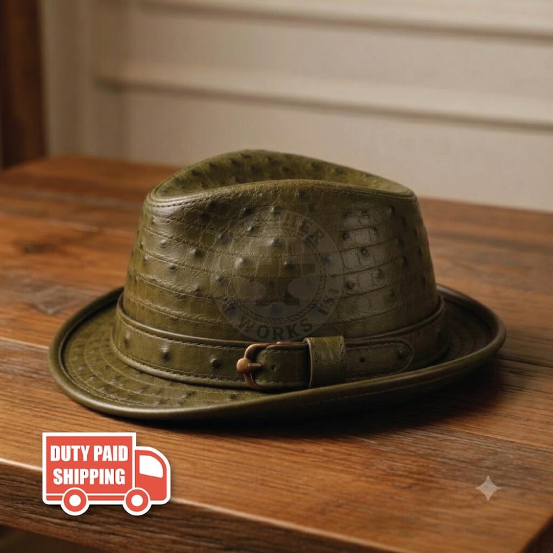 May include: An olive green fedora hat featuring a textured ostrich-skin pattern across the crown and brim. The hat includes a matching olive green band around the base, secured with a small, metallic buckle on the side for a structured appearance.