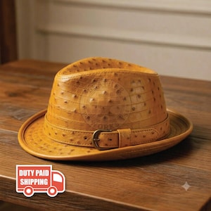 May include: A tan fedora hat featuring a textured ostrich-skin pattern across the crown and brim. The hat includes a matching tan band around the base, secured with a metallic buckle on the side, creating a structured and classic headwear piece.