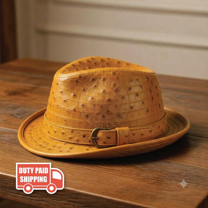 May include: A tan fedora hat featuring a textured ostrich-skin pattern across the crown and brim. The hat includes a matching tan band around the base, secured with a metallic buckle on the side, creating a structured and classic headwear piece.