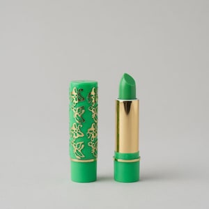 May include: Green lipstick with a gold-colored base and a green cap. The cap is decorated with a gold butterfly pattern. The lipstick is open, revealing the green color of the lipstick.