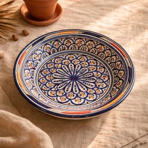 Könnte beinhalten: Eine runde Keramikschale mit einem detaillierten, handbemalten Muster in den Farben Dunkelblau, Orange und Schwarz. Das Design besteht aus einem zentralen, blumenartigen Mandala-Motiv, das von konzentrischen Ringen aus geometrischen Bögen umgeben ist.