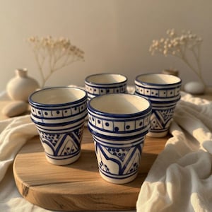 Peut inclure: Quatre tasses en céramique blanche avec des motifs géométriques bleus. Les tasses ont une forme cylindrique avec un bord légèrement évasé. Le motif bleu comprend des lignes, des points et des formes triangulaires. Les tasses sont disposées sur une surface en bois.