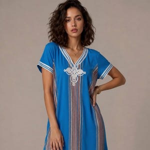 Könnte beinhalten: Ein leuchtend blaues Kaftankleid mit V-Ausschnitt, weißer Zierleiste und einem zentralen, weiß bestickten Blumenmotiv. Das Kleidungsstück hat kurze Ärmel mit weiß gestreiften Bündchen sowie vertikale, mehrfarbige Streifen in Braun und Weiß.