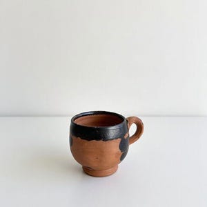 Peut inclure: Une petite tasse en terre cuite faite à la main, avec un corps arrondi et une petite anse. Le bord et une partie de l'extérieur sont peints avec des accents noirs. La tasse a un aspect rustique et artisanal.