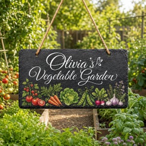 May include: A rectangular black slate sign hanging from a brown rope, featuring the white text "Olivia's Vegetable Garden" above a colorful illustration of tomatoes, carrots, lettuce, peas, radishes, and garlic bulbs on a dark background.
