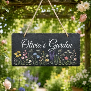 Pode incluir: Uma placa retangular de ardósia preta pendurada por uma corda de juta. A placa apresenta o texto branco Olivia's Garden em fonte cursiva, posicionado acima de uma ilustração colorida de várias flores silvestres em tons de amarelo, rosa, azul e roxo.