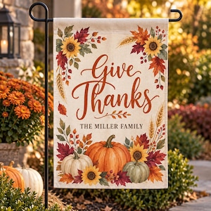以下が含まれることがあります： Give ThanksおよびThe Miller Familyという文字が茶色の筆記体とブロック体で記された長方形のガーデンフラッグ。オレンジと白のカボチャ、黄色のヒマワリ、赤と金の秋の葉のイラストが描かれています。