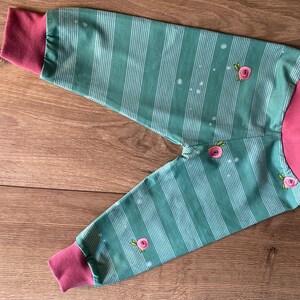 Puede incluir: Pantalones de bebé a rayas verdes y blancas, con puños y cintura rosas. El diseño incluye pequeños bordados de rosas rosas y motas blancas. Los pantalones de tejido suave están diseñados para la comodidad.
