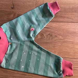 Puede incluir: Prenda de manga larga a rayas verdes y blancas con puños rosas. La tela presenta pequeños lunares blancos y una pequeña flor bordada. La prenda es de material suave y es adecuada para niños.