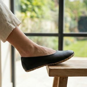 May include: A black flat shoe with a rounded toe and a visible light-colored stitched sole edge. The shoe features a simple, low-profile design with a smooth upper surface and a reinforced heel counter, resting on a wooden surface.