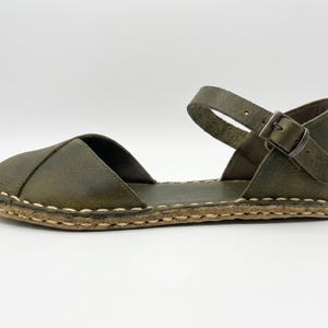 Peut inclure: Une sandale en cuir vert olive avec un bout ferm&eacute; arrondi, un c&ocirc;t&eacute; ouvert et une bride de cheville fix&eacute;e par une boucle en m&eacute;tal. La semelle pr&eacute;sente une bordure &eacute;paisse en jute tress&eacute; de couleur claire avec des coutures apparentes.