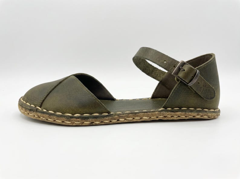 Peut inclure: Une sandale en cuir vert olive avec un bout ferm&eacute; arrondi, un c&ocirc;t&eacute; ouvert et une bride de cheville fix&eacute;e par une boucle en m&eacute;tal. La semelle pr&eacute;sente une bordure &eacute;paisse en jute tress&eacute; de couleur claire avec des coutures apparentes.