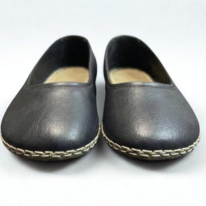 May include: A pair of black slip-on flats featuring a rounded toe and a visible light-colored whipstitch detail along the outer edge of the sole. The interior of the shoes is lined with a light beige material, and the exterior has a smooth finish.