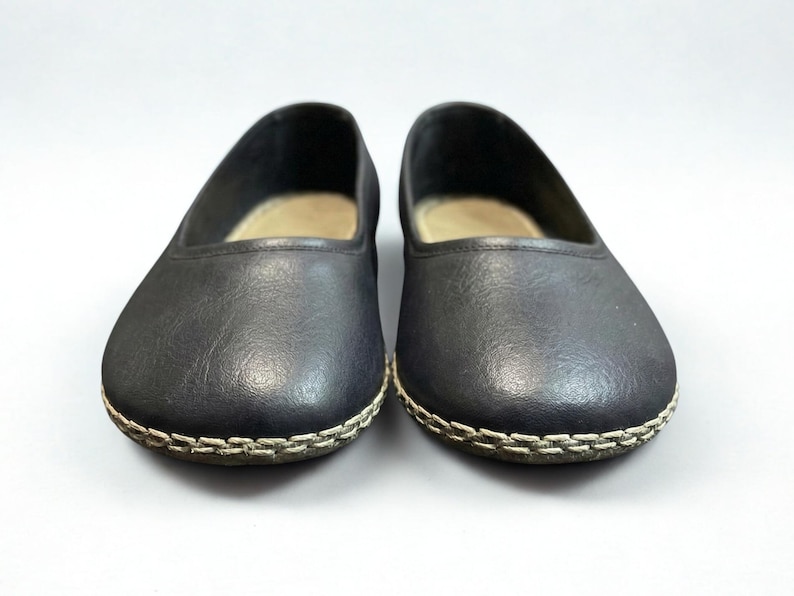 May include: A pair of black slip-on flats featuring a rounded toe and a visible light-colored whipstitch detail along the outer edge of the sole. The interior of the shoes is lined with a light beige material, and the exterior has a smooth finish.