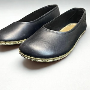 May include: A pair of black slip-on flats featuring a rounded toe and a smooth leather-like upper. The shoes are constructed with a visible light-colored whipstitch detail along the sole edge and include a tan interior insole.