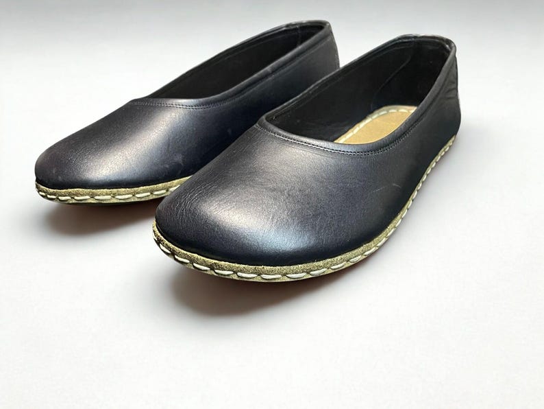 May include: A pair of black slip-on flats featuring a rounded toe and a smooth leather-like upper. The shoes are constructed with a visible light-colored whipstitch detail along the sole edge and include a tan interior insole.