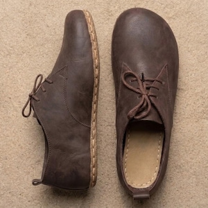 Puede incluir: Un par de zapatos de cuero marrón oscuro con puntera ancha y redondeada y cierre de cordones. Los zapatos presentan costuras visibles de color claro a lo largo del borde de la suela, un diseño de perfil bajo y una pequeña trabilla en el talón.