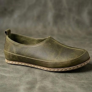 May include: An olive green slip-on shoe featuring a smooth leather upper, a rounded toe, and a visible light-colored stitched welt connecting the upper to the sole. A small pull tab is located at the heel of the shoe, which has a low profile.