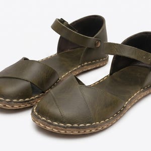 Peut inclure: Une paire de sandales plates vert olive dot&eacute;es d'un design &agrave; bout ferm&eacute; avec des brides crois&eacute;es, une bride de cheville avec une boucle en m&eacute;tal et des coutures claires visibles le long de la semelle. Les sandales ont une finition mate.