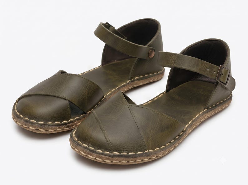 Peut inclure: Une paire de sandales plates vert olive dot&eacute;es d'un design &agrave; bout ferm&eacute; avec des brides crois&eacute;es, une bride de cheville avec une boucle en m&eacute;tal et des coutures claires visibles le long de la semelle. Les sandales ont une finition mate.