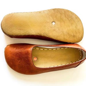 May include: A pair of brown leather slip-on shoes featuring a rounded toe, a smooth tan interior lining, and visible white stitching along the collar. The bottom shoe shows a textured tan sole with a small circular metal rivet embedded in the center.
