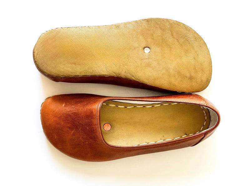May include: A pair of brown leather slip-on shoes featuring a rounded toe, a smooth tan interior lining, and visible white stitching along the collar. The bottom shoe shows a textured tan sole with a small circular metal rivet embedded in the center.