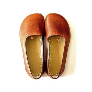 May include: A pair of brown leather slip-on shoes. The shoes have a rounded toe and a flat sole. The interior is a lighter tan color, with visible stitching along the edges. A small copper-colored rivet is visible on the top of each shoe.