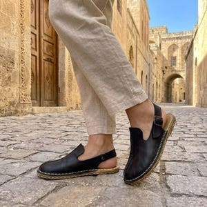 May include: Black leather sandals with a cork sole and a buckle closure. The sandals have a slip-on design with an open back and a stitched edge. The person is wearing beige pants.