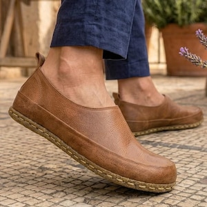 Puede incluir: Zapatos sin cordones de cuero marr&oacute;n con punta redondeada y suela cosida. Los zapatos se usan con pantalones azul marino. La imagen est&aacute; tomada al aire libre en un patio de piedra. Los zapatos parecen c&oacute;modos e informales.