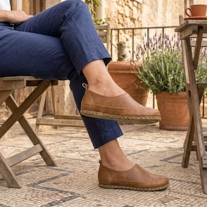 Puede incluir: Zapatos marrones de cuero sin cordones con suela cosida. Los zapatos se llevan con pantalones azul marino. La imagen est&aacute; tomada al aire libre, con una silla y una mesa de madera en el fondo.