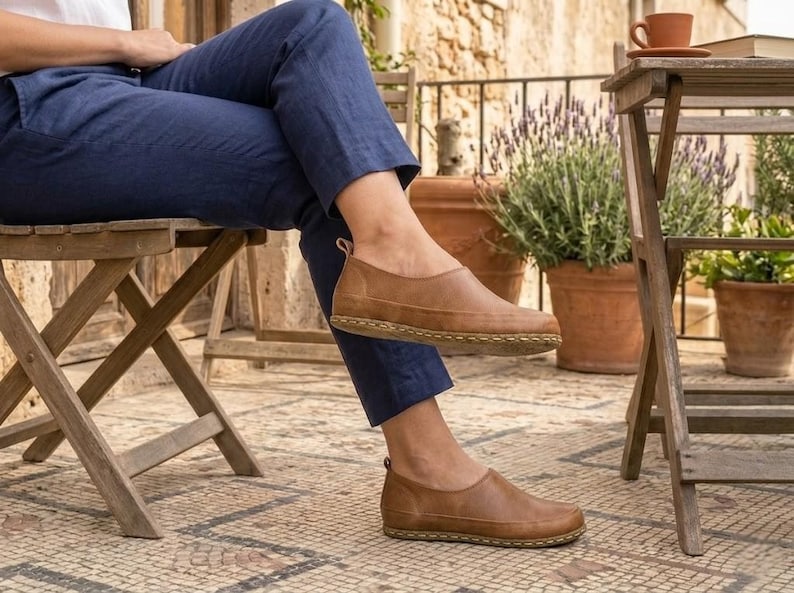 Puede incluir: Zapatos marrones de cuero sin cordones con suela cosida. Los zapatos se llevan con pantalones azul marino. La imagen est&aacute; tomada al aire libre, con una silla y una mesa de madera en el fondo.