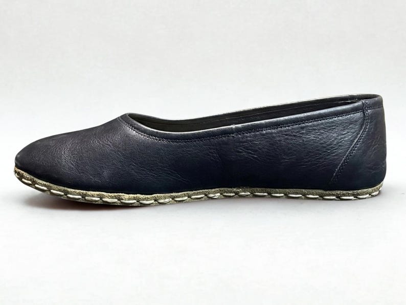 May include: A dark navy blue leather flat shoe with a rounded toe and a low-cut topline. The shoe features a visible, thick white whipstitch detail along the entire perimeter where the upper leather meets the sole, creating a distinct contrast.
