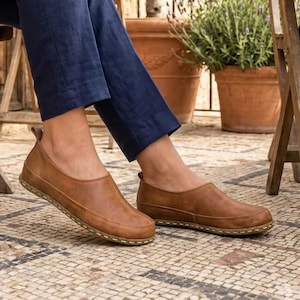 Puede incluir: Zapatos sin cordones de cuero marr&oacute;n con punta redondeada y suela cosida. Los zapatos se usan con pantalones azules. El fondo incluye una silla de madera y plantas en macetas. Los zapatos son de cuero.
