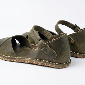 Peut inclure: Une paire de sandales en cuir vert olive dot&eacute;es d'une large bride sur le dessus, d'une bride de cheville r&eacute;glable avec une boucle en m&eacute;tal et d'un contrefort ferm&eacute;. La semelle est fix&eacute;e &agrave; la tige par des coutures apparentes de couleur claire.
