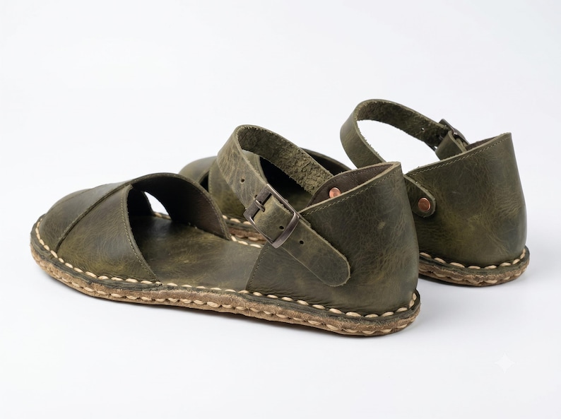 Peut inclure: Une paire de sandales en cuir vert olive dot&eacute;es d'une large bride sur le dessus, d'une bride de cheville r&eacute;glable avec une boucle en m&eacute;tal et d'un contrefort ferm&eacute;. La semelle est fix&eacute;e &agrave; la tige par des coutures apparentes de couleur claire.