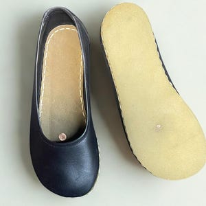 May include: A pair of dark navy blue slip-on shoes featuring a smooth leather upper, a rounded toe, and a light beige textured sole. The interior insole is light beige with visible white stitching along the edge and a small circular metallic detail.