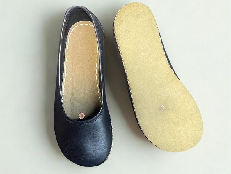 May include: A pair of dark navy blue slip-on shoes featuring a smooth leather upper, a rounded toe, and a light beige textured sole. The interior insole is light beige with visible white stitching along the edge and a small circular metallic detail.