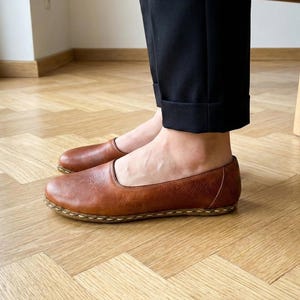 May include: A pair of brown leather slip-on shoes featuring a rounded toe, a low-cut topline, and visible light-colored stitching along the sole edge. The shoes have a smooth, matte finish and are worn with black trousers.