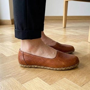 May include: A pair of brown leather slip-on flats featuring a rounded toe, a low-cut topline, and visible light-colored stitching along the sole edge. The shoes are worn with black cuffed trousers, showcasing a smooth, textured finish on the leather.