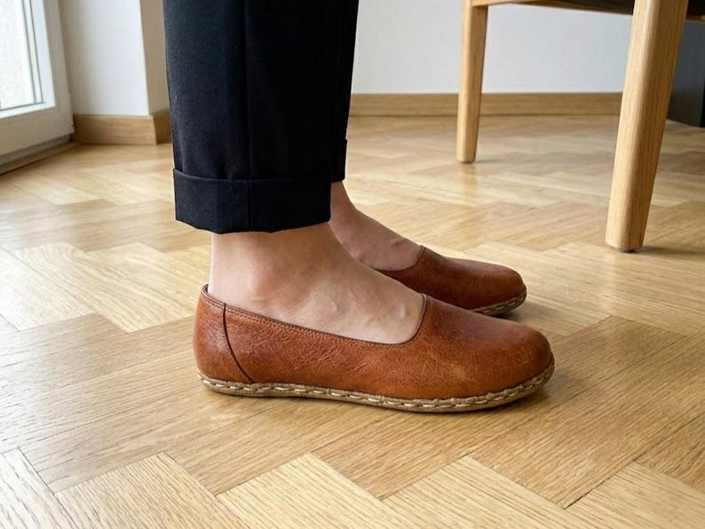 May include: A pair of brown leather slip-on flats featuring a rounded toe, a low-cut topline, and visible light-colored stitching along the sole edge. The shoes are worn with black cuffed trousers, showcasing a smooth, textured finish on the leather.