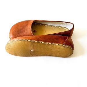 May include: A pair of tan leather slip-on shoes featuring a rounded toe, a smooth upper surface, and a light-colored, textured sole attached with visible thick stitching. The interior lining is a light cream color with a small circular cutout.